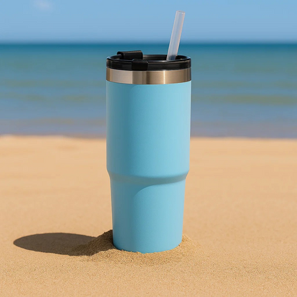 Blue tumbler with a black lid and straw on a sandy beach with ocean in the background