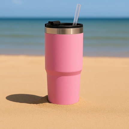 Pink tumbler with a straw on a sandy beach with ocean in the background